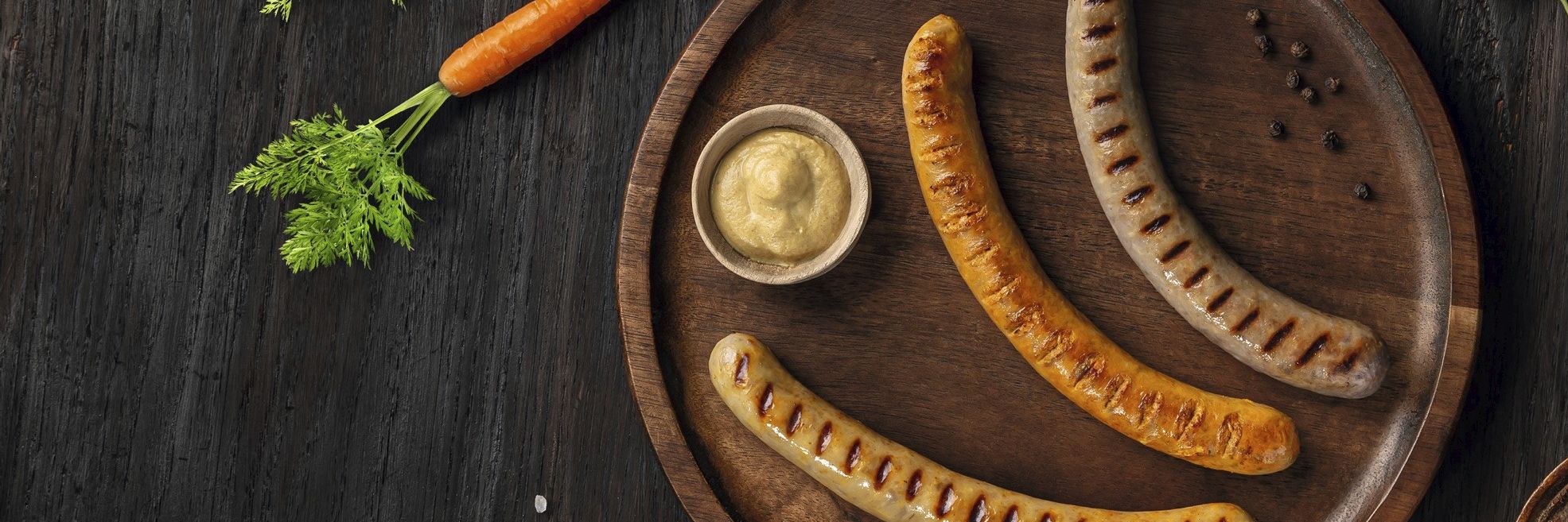 Drei hybride Bratwurstvarianten – mit Karotte, Mais und Rotkohl – auf einem Brett vor dunklem Hintergrund. Beispiel für moderne Hybridprodukte für Metzgereien, Lebensmittelindustrie und Systemgastronomie.