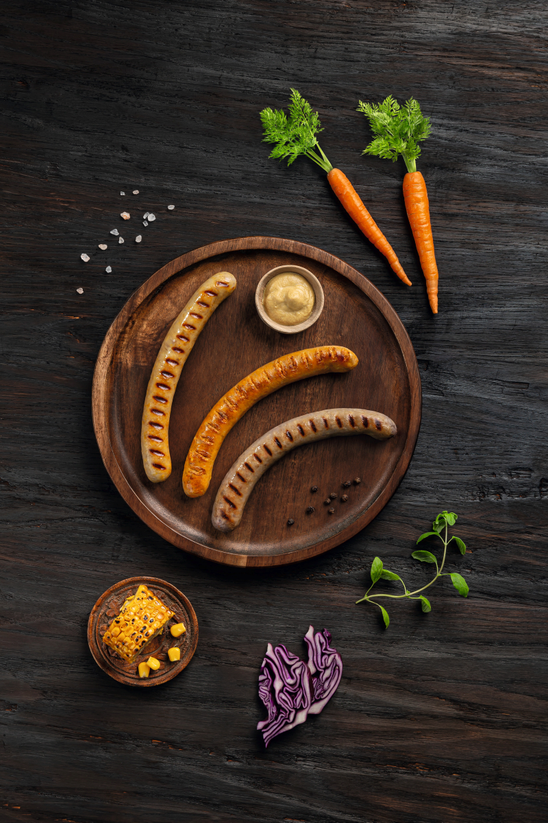 Hochformatige Darstellung von drei hybriden Bratwürsten mit Karotte, Mais und Rotkohl auf einem Brett vor dunklem Hintergrund – innovative Hybridprodukte für Metzgereien, Lebensmittelindustrie und Systemgastronomie.