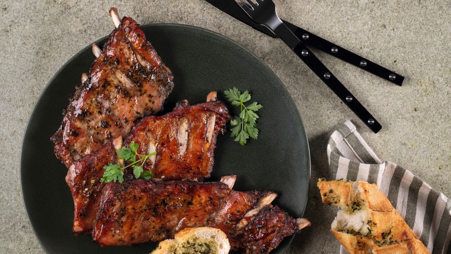 Glasierte Spareribs mit Kräuterbaguette, Dip und Krautsalat auf einer dunklen Platte