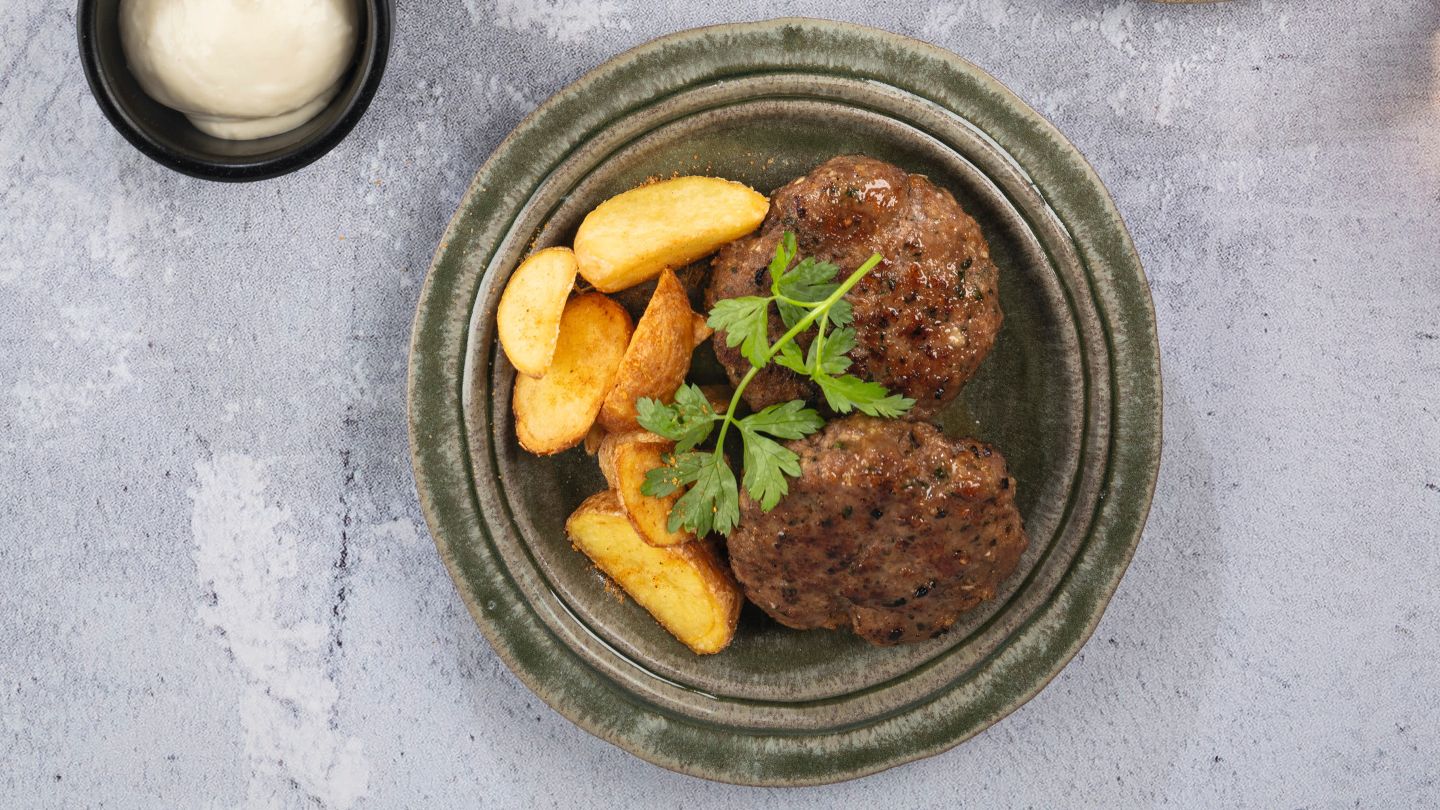 Frikadellen im Airfryer-Korb und in zwei Schalen mit Kartoffelbeilage, Gemüse und verschiedenen Saucen