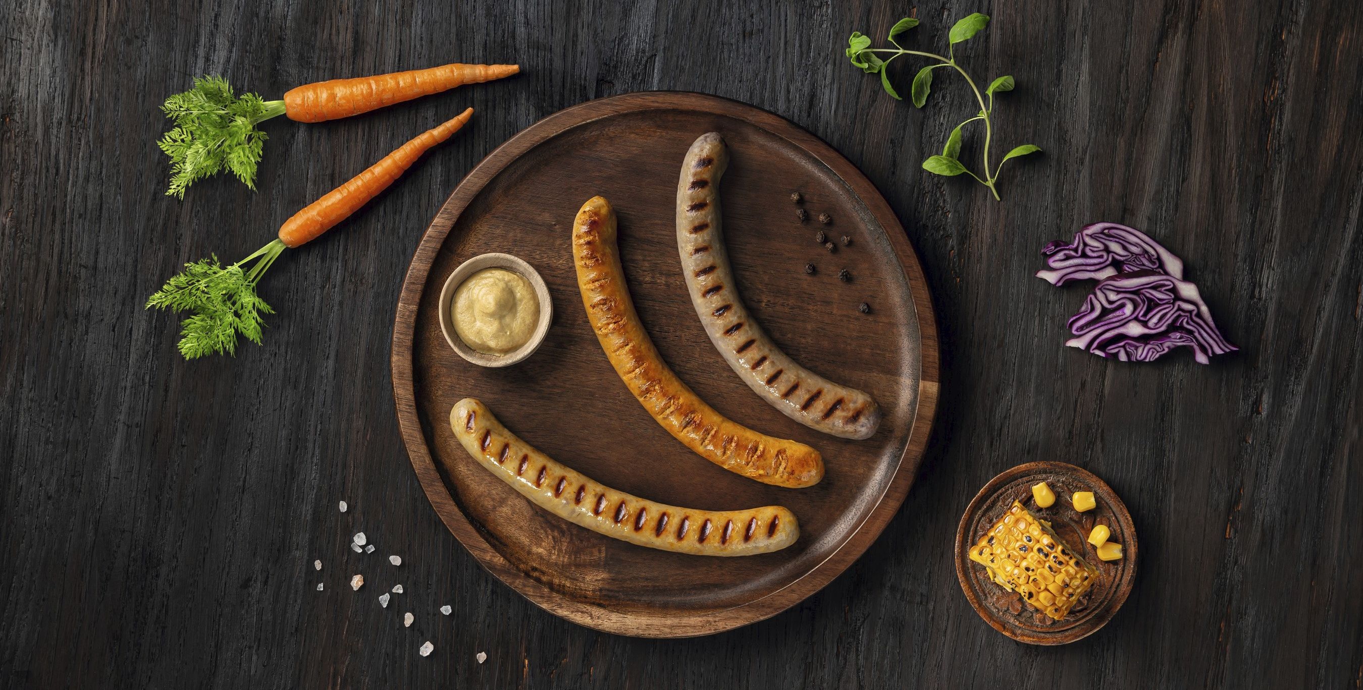 Drei hybride Bratwurstvarianten – mit Karotte, Mais und Rotkohl – auf einem Brett vor dunklem Hintergrund. Beispiel für moderne Hybridprodukte für Metzgereien, Lebensmittelindustrie und Systemgastronomie.